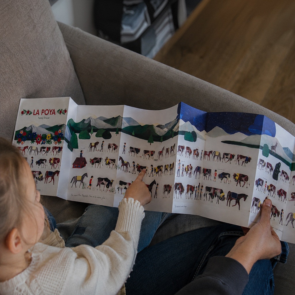 Child and adult reading a long folding illustrated poster showing horses in a mountainous landscape indoors on a sofa.