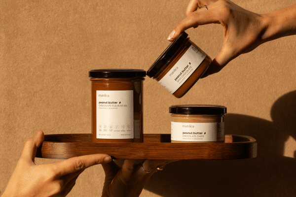 Three jars of peanut butter arranged on a wooden serving tray, with a hand lifting one jar to display its label.