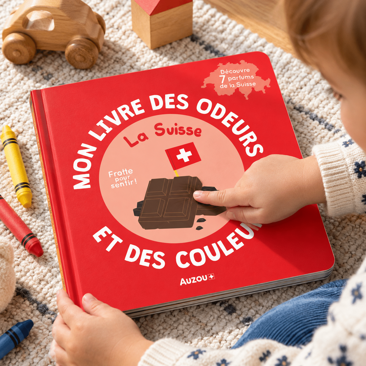 Child's hands hold a bright red children's book titled 'Mon Livre des Odeurs et des Couleurs' on a textured rug, with toy blocks and crayons nearby.