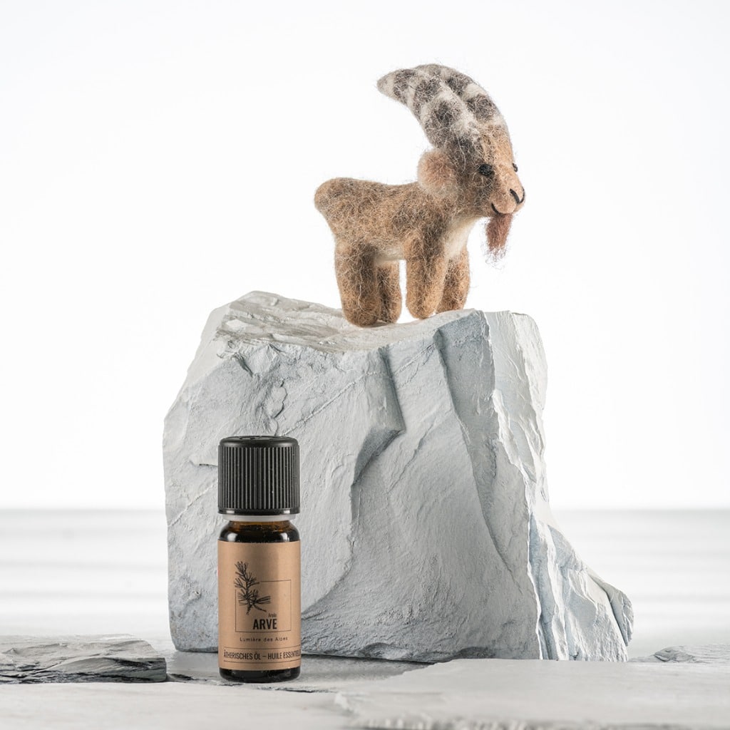 Amber glass bottle of Arve essential oil with a black cap, set on a pale gray rock with a woolly goat figurine perched atop it.
