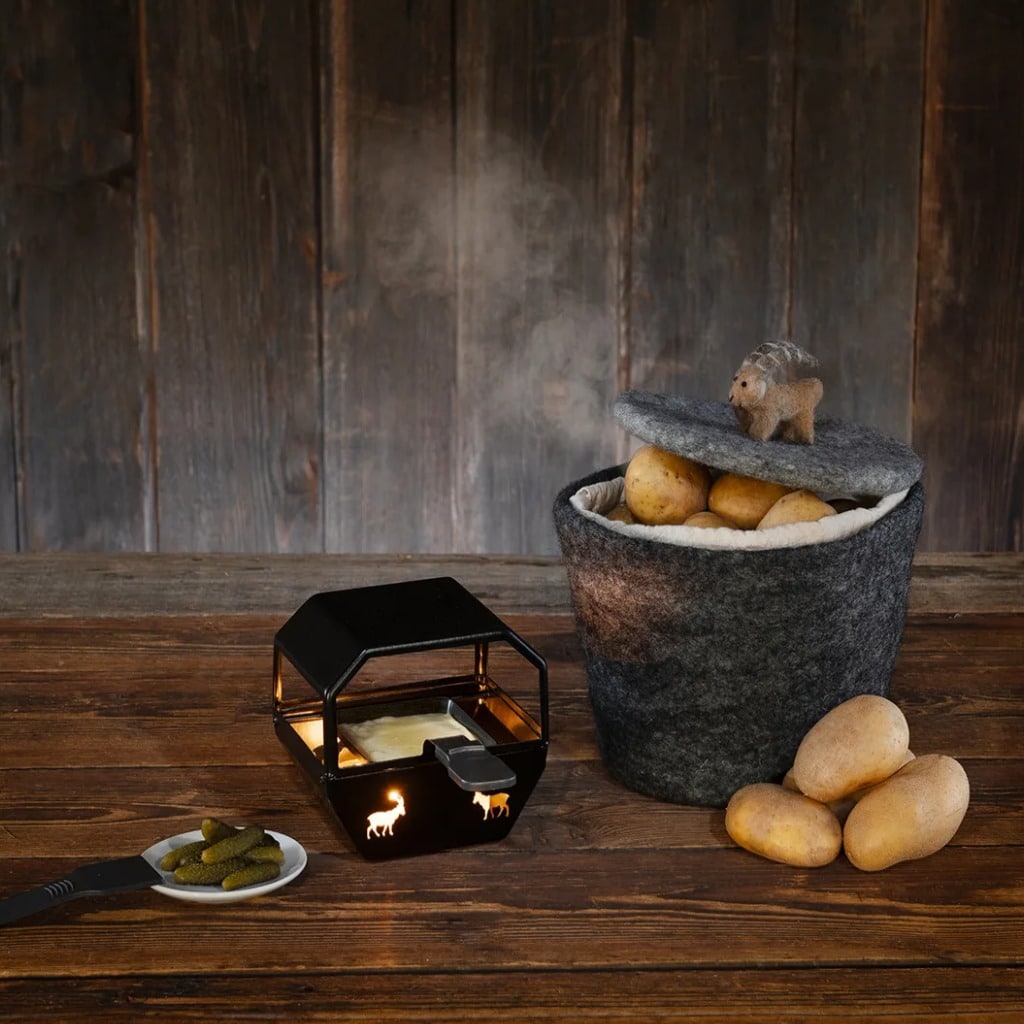Sauna scene with a black stove emitting a warm glow, a felted bucket of potatoes with a lid and a small animal figurine on top, plus a plate of pickles and a few potatoes on a wooden table.